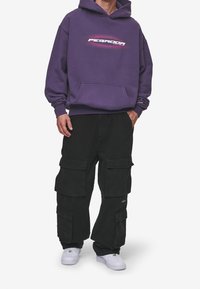 Fialová mikina s kapucí a logem na hrudi, doplněná černými cargo kalhotami s několika kapsami a bílými teniskami.