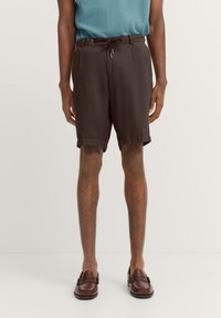Dunkelbraune Shorts aus leichtem Material mit einem Kordelzugbund, entspannter Passform und seitlichen Taschen. Getragen mit braunen Slippern.