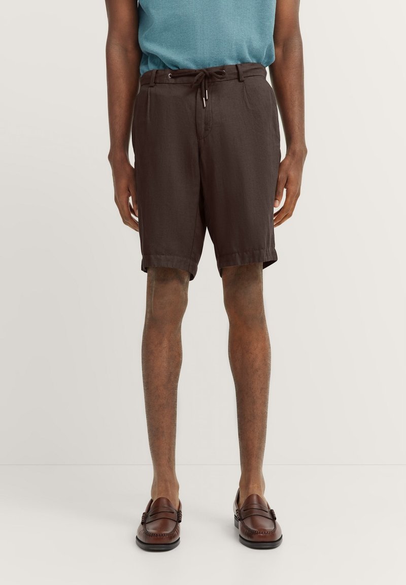 Dunkelbraune Shorts aus leichtem Material mit einem Kordelzugbund, entspannter Passform und seitlichen Taschen. Getragen mit braunen Slippern.