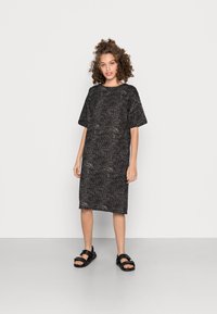 Vestido preto com um corte solto, mangas curtas e um decote redondo. Apresenta um padrão sutil salpicado. Combinado com sandálias planas pretas.