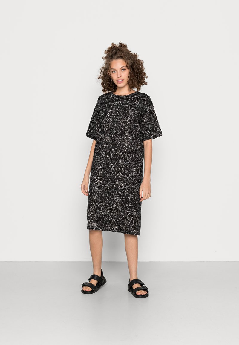 Vestido preto com um corte solto, mangas curtas e um decote redondo. Apresenta um padrão sutil salpicado. Combinado com sandálias planas pretas.