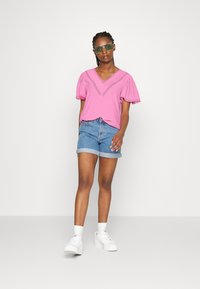 Top rosa de mangas curtas com decote em V e ornamentos decorativos, combinado com calções de ganga enrolados e ténis brancos. Fundo simples.