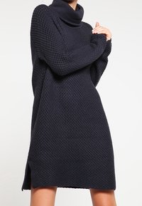 Robe tricotée bleu marine avec un motif texturé, col roulé, manches longues et fentes latérales ; fabriquée dans un matériau épais et confortable.