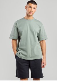 Green cotton t-shirt with a crew neck, short sleeves, relaxed fit, paired with black shorts. Smooth texture, no visible patterns or accents.