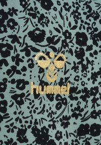 Tkanina w kwiatowy wzór w kolorze zielonym z czarnymi kwiatami. Na dole, na środku, znajduje się złoty błyszczący logo "hummel". Materiał o fakturze dzianiny.