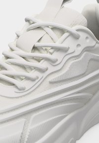 Gros plan d'une chaussure de sport blanche montrant les lacets, le tissu en maille texturé et les détails de la semelle en caoutchouc sculptée.