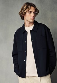 Schwarze strukturiertes Jacke mit zwei vorderen Taschen, Druckknöpfen und einem spitzen Kragen, getragen über einem hellen Hemd. Einfaches Design, lässige Passform.