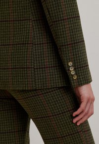 Groen geruit pak met een gestructureerde wolmix, met kastanjebruine en marineblauwe lijnen. Drie goudkleurige knopen als detail bij de manchetten.