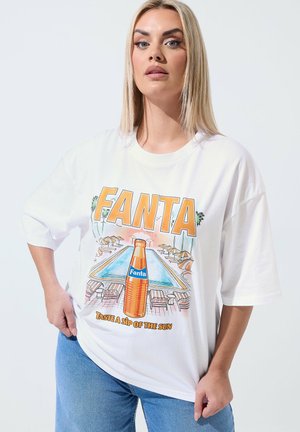 Kvinde med glat blondt hår iført en hvid oversized T-shirt med en Fanta-flaske, stående ved en pool med stole og parasoller, kombineret med blå jeans.