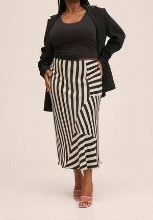 Kvinde iført sort top, sort blazer, sort og hvid verticalstribet midinederdel og lyserøde åbne sko med hæl, stående med hænderne på hofterne.