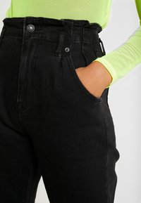 Zwarte high-waisted denim broek met een knoop- en ritsluiting, voorzien van zijzakken en een gestructureerde afwerking. De tailleband heeft riemlussen.