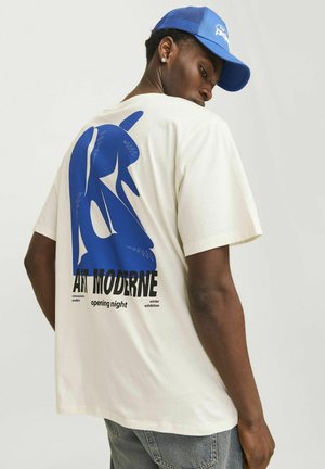 T-shirt blanc avec un grand motif graphique bleu au dos accompagné de texte. Porté avec une casquette bleue et un jean en denim.