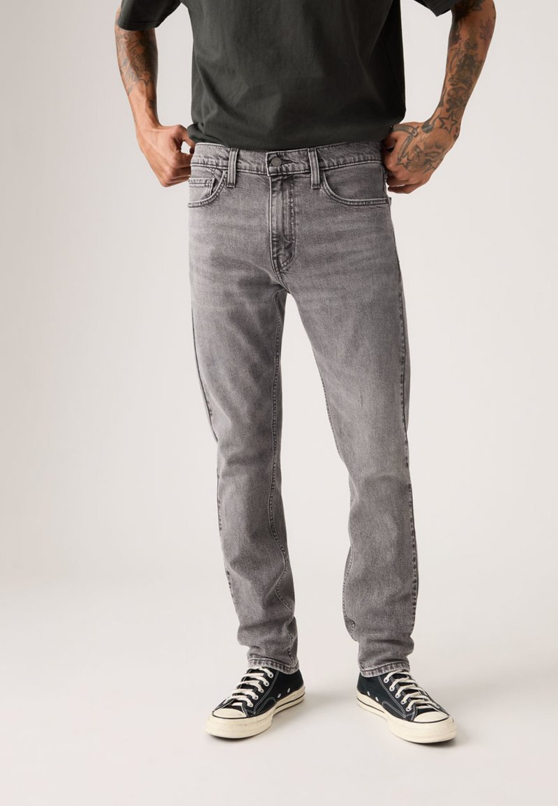 Jeans de mezclilla gris con diseño de corte recto, estilo de cinco bolsillos y desvanecimiento ligero. Combinados con zapatillas negras bajas.