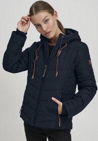 Veste matelassée marine avec capuche, présentant des accents en cuir marron et une fermeture éclair. Des coutures verticales ajoutent de la texture.