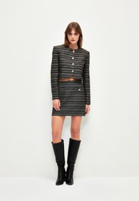 Chaqueta de tweed recortada en negro y dorado con botones redondos, combinada con una falda ajustada a juego. Acompañada de botas negras hasta la rodilla.