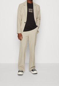 Beige rutigt kostym med en strukturerad blazer och lösa byxor, ihop med en svart t-shirt med vit logotext och svarta sneakers.