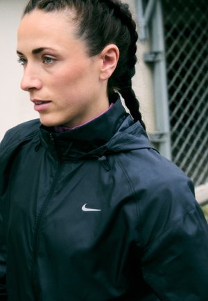Veste Nike noire avec un col haut et un logo discret, fabriquée dans un tissu léger et brillant, dotée de cordons de serrage réglables au niveau du cou.