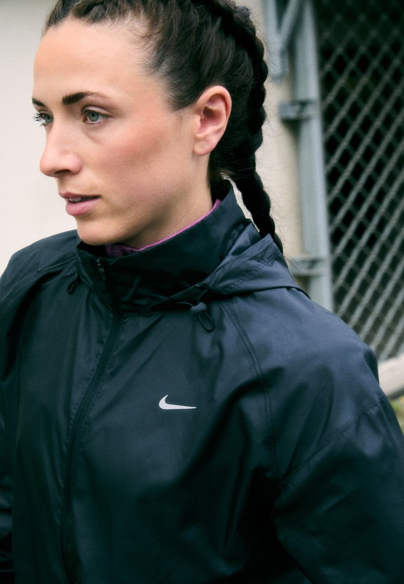 Sort Nike-jakke med højt krave og subtile logoer, fremstillet af let, skinnende stof og justerbare snore ved halsen.