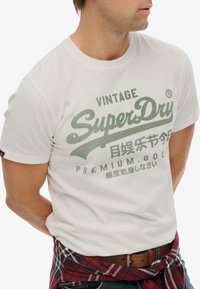 Camiseta de algodón blanca con gráfico verde "SuperDry". Mangas cortas y cuello redondo. Una camisa de cuadros atada a la cintura añade contraste.