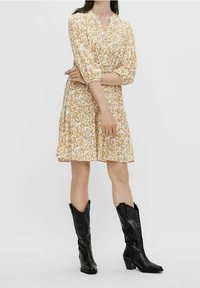 Florales Kleid aus gelbem und weißem Stoff mit 3/4-Ärmeln, V-Ausschnitt und weit ausgestelltem Saum. Kombiniert mit glänzenden schwarzen Cowboy-Stiefeln.