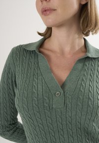 Pull en maille torsadée vert avec un col en V, doté d'une patte de boutonnage à deux boutons. Texture douce et motif en maille détaillé sur l'ensemble.