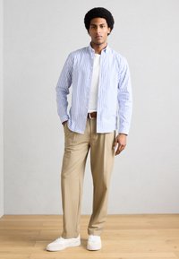 Νέος άνδρας που στέκεται με τα χέρια στις τσέπες, φορώντας λευκά αθλητικά παπούτσια, μπεζ παντελόνι, λευκό μπλουζάκι και μπλε-λευκό ριγέ πουκάμισο με κουμπιά.