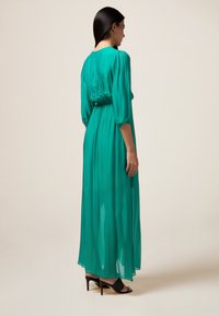 Rochie lungă, fluidă, dintr-un material teal vibrant. Prezintă o talie strânsă, mâneci până la trei sferturi și o textură moale, transparentă. Purtați-o cu sandale negre cu toc.