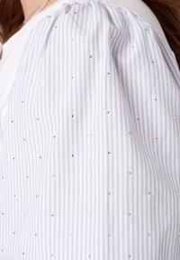 Blouse en tissu rayé avec des rayures verticales blanches et bleu clair, dotée de petits embellissements brillants répartis uniformément sur le tissu.