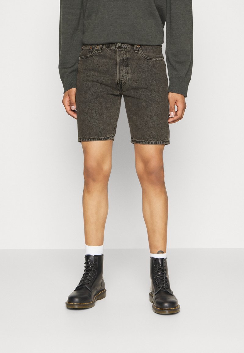 Shorts en denim noir à coupe droite, texture délavée et coutures contrastantes. Associés à des bottes noires à lacets et des chaussettes blanches.