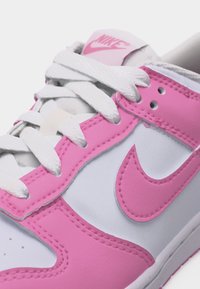 Pantof sport Nike alb și roz, cu șireturi albe, logo swoosh roz și detalii de cusături roz.