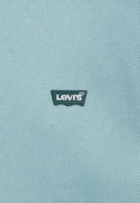 Um tecido de malha azul claro com um logótipo da Levi's bordado em verde escuro no canto inferior esquerdo, exibindo um design de etiqueta de forma clássica.