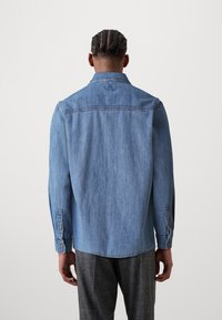 Blaue Denim-Hemd mit langen Ärmeln, lässigem Kragen, Knopfakzenten an den Manschetten und leicht strukturiertem Finish. Die Rückansicht zeigt die Yoke-Details.