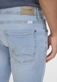 Jeans din denim de culoare albastru deschis, cu o finisare texturată, având o buzunare pe spate, cusături vizibile și un eticheta din piele maro a brandului.