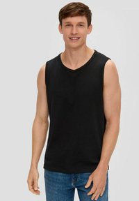 Young man smiling, wearing a sleeveless black shirt and blue jeans, standing against a plain light background.