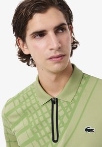 Camisa polo verde con un patrón geométrico, cuello corto con cremallera, collar acanalado y el logo del cocodrilo de Lacoste en el pecho. Tejido texturizado.
