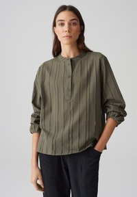 Camicia verde oliva con bottoni, scollo rotondo, maniche lunghe e righe verticali. Realizzata in tessuto leggero e testurizzato.