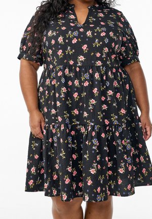 Frau trägt ein schwarzes Kleid mit rosa und blauen Blumenmustern und kurzen Puffärmeln vor weißem Hintergrund.