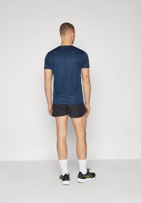 Tumed sinine spordit-särk ja mustad lühikesed püksid, sujuva tekstuuriga; inimene on nähtav seljapoolelt, jalas mustad kingad roheliste aksessuaaridega.