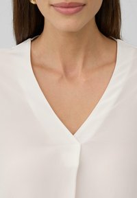 Biała bluzka z dekoltem w serek, wykonana z gładkiego, lekkiego materiału. Minimalistyczny design, bez widocznych wzorów czy ozdób.