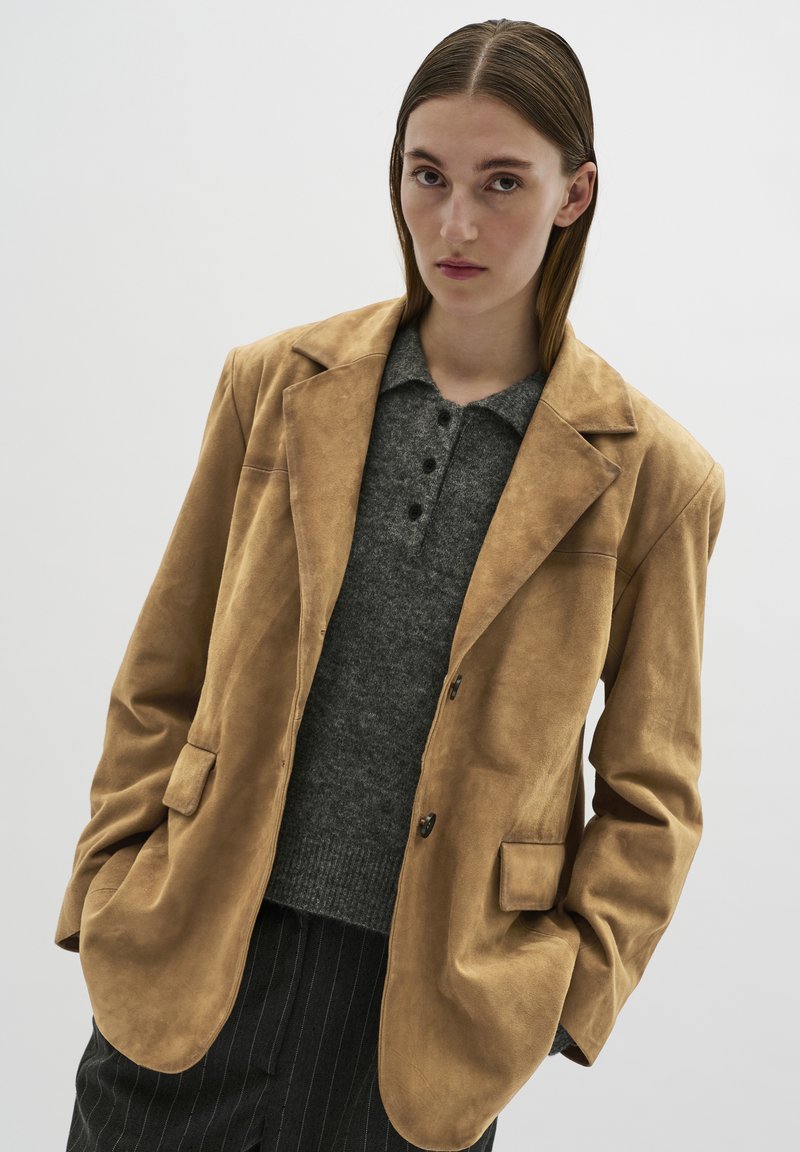 Tan suède blazer met Engelse revers en twee voorkwabben, gelaagd over een charcoal grijs gemêleerd poloshirt. Verticale gestreepte broek zichtbaar.