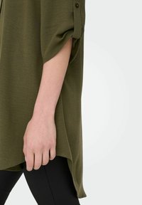Camicia verde oliva ample con maniche arrotolate, tessuto texturizzato e orlo asimmetrico, abbinata a leggings neri.