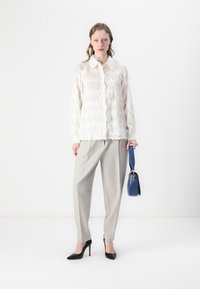 Blusa bianca con texture e motivo a zigzag, pantaloni grigi chiari a vita alta, tacchi neri a punta e una borsa blu. Design semplice.