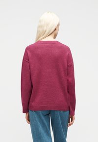 Γυναίκα με μακριά ξανθά μαλλιά που φορά χαλαρό μωβ πουλόβερ και μπλε τζιν, στέκεται με την πλάτη προς την κάμερα.