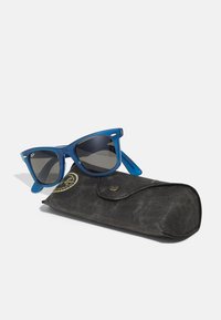 Blå plastsolbriller med mørke linser, som har en klassisk fasong og diskret Ray-Ban-merkevare, ligger ved siden av et svart teksturert etui.