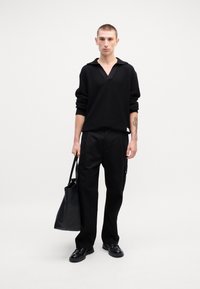 Schwarzer Strickpullover mit V-Ausschnitt und langen Ärmeln, kombiniert mit schwarzen Hosen. Das Modell trägt eine schwarze Leder-Tote-Bag und schwarze Schuhe.