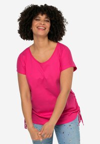 Camiseta de algodón rosa brillante con escote redondeado, mangas cortas y lazos ajustables a los lados. Combinada con jeans azul claro que presentan adornos coloridos.