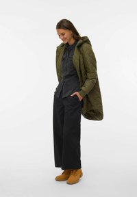 Zelena prešita jakna s kapuco, temna srajca z gumbi, črne široke hlače in rjavi gležnarji. Gladke teksture in večplastna zasnova.