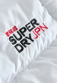 Tessuto bianco con trama trapuntata che presenta la scritta ricamata "SUPER DRY" in nero e "JPN" in rosso, arricchita da caratteri asiatici sopra il logo.