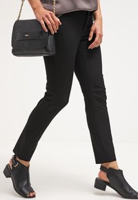 Černé slim-fit kalhoty vyrobené z hladké tkaniny, doplněné otevřenými černými lodičkami s mohutným podpatkem. Malá černá kabelka s řetízkovým popruhem.