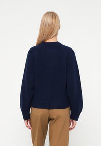 Blondīne, kas tērpusies tumši zilā adītā džemperī un gaišās biksēs, stāv ar muguru pret vienkāršu baltu fonu.